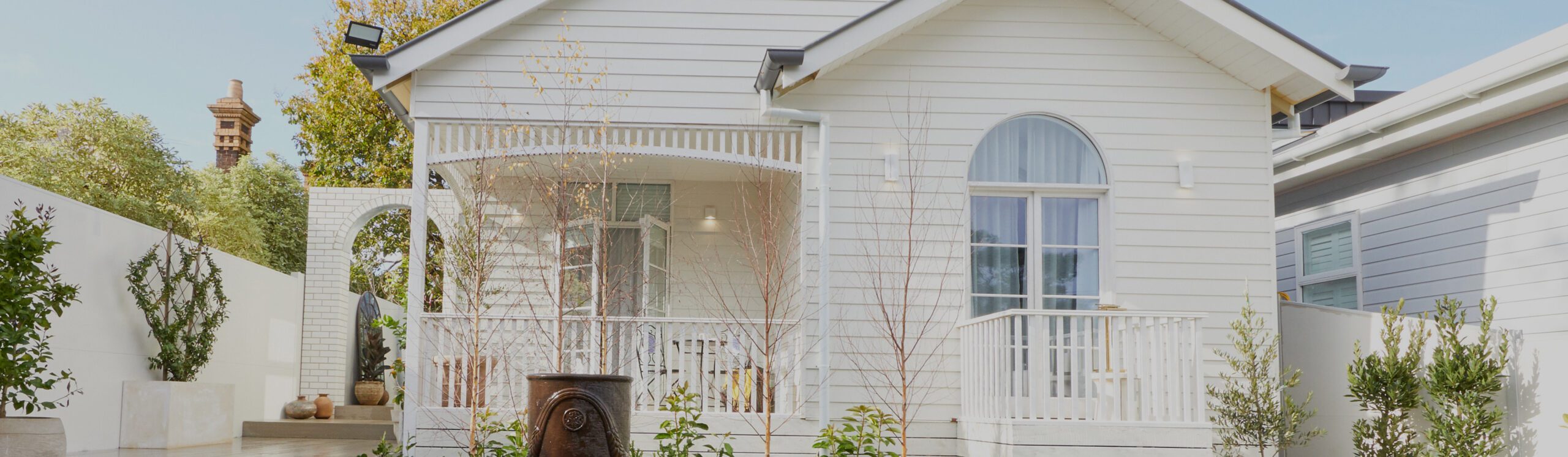 A charming white house with a veranda adorned with latticework. The front yard features various potted plants and a small water fountain. The home has a large arched window and is set against a backdrop of greenery and a bright blue sky.