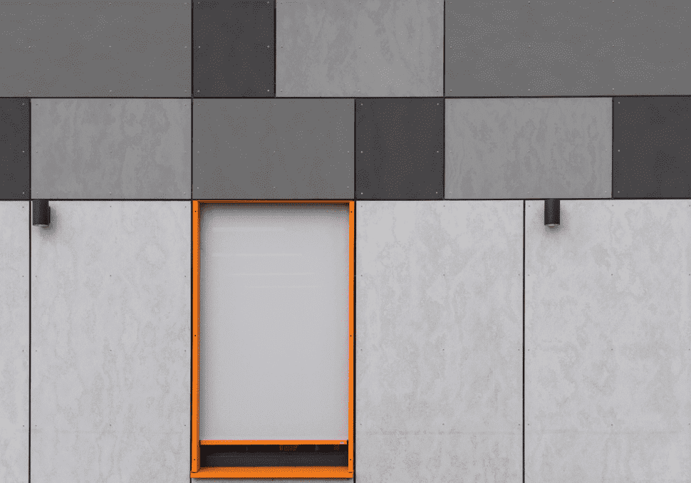 A minimalist grey building facade with a mix of dark and light rectangular panels. In the center is a window with an orange frame, partially covered by a blind. Two small wall-mounted lights are symmetrically placed on either side of the window.