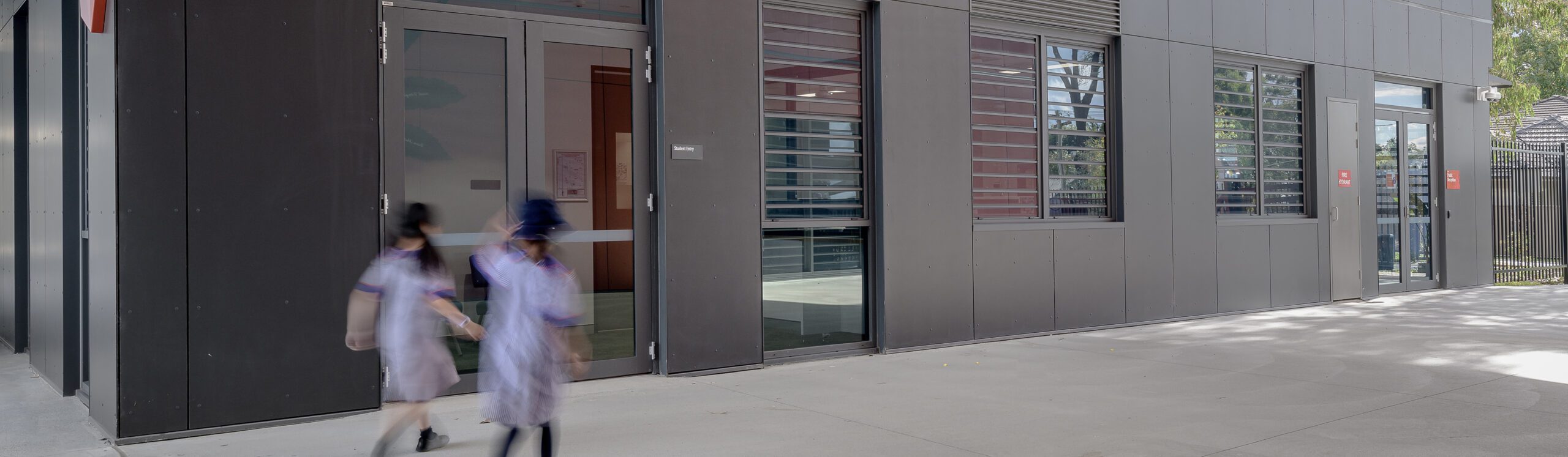 Two children wearing school uniforms walk along the sidewalk next to a modern building with dark panels, large windows, and glass doors. The scene appears to be during the day with some sunlight and shadows visible.